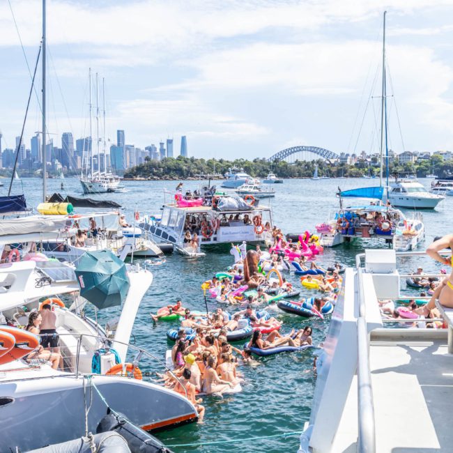 A group of people on boats and inflatables gather in Sydney Harbour, enjoying a sunny day with the city skyline and Sydney Harbour Bridge in the background, complemented by a luxurious private yacht charter.