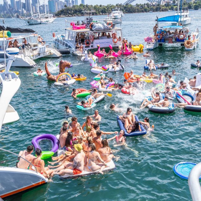 Numerous people are swimming and lounging on inflatable floats in a busy harbor, with boats anchored nearby and a city skyline, including a distinctive arched bridge, visible in the background. Consider a private yacht charter Sydney Harbour for an unforgettable experience amidst this vibrant setting.