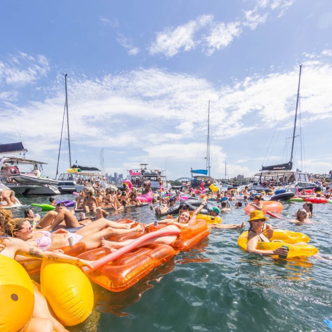 Description: People relaxing on large inflatable floats in the water surrounded by boats, under a bright blue sky, enjoying a private yacht charter on Sydney Harbour.