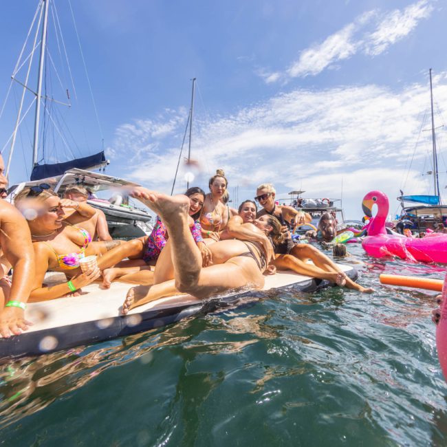 A group of people in swimwear enjoying a sunny day on the water with boats, floaties, and water noodles around them during a luxury yacht hire Sydney experience.