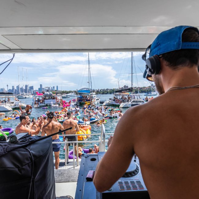 A DJ in a blue cap performs on a catamaran while people on nearby boats and inflatables enjoy the water on a sunny day. The city skyline is visible in the background.