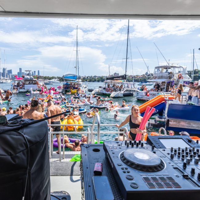 A DJ booth overlooks a crowded boat party on the water with numerous people swimming and floating; boats and a city skyline appear in the background. It’s the ultimate vibe for corporate boat events Sydney, offering an unmatched party atmosphere.