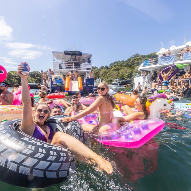 A group of people enjoy a sunny day on the water, floating on various inflatables including a unicorn, flamingo, and tire, with boats and more people visible in the background. A DJ boat hire from Sydney adds lively music to the cheerful scene.