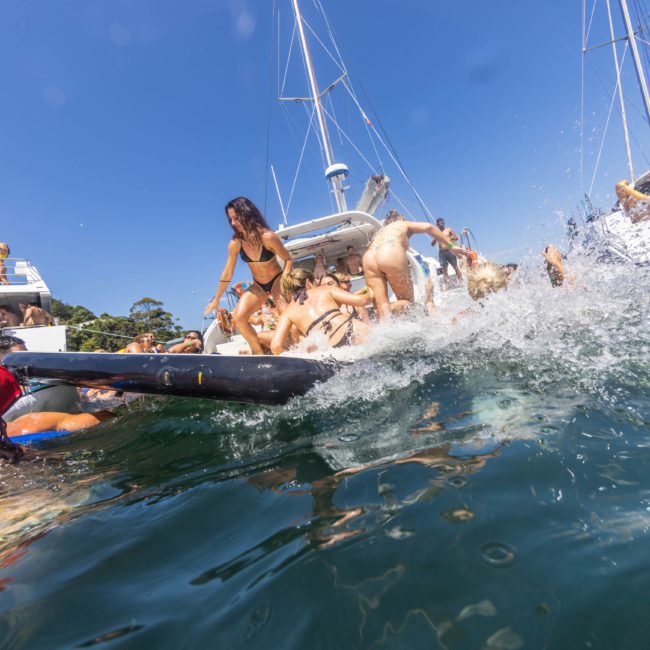 People enjoying an active summer party on a private yacht charter in Sydney Harbour, with several individuals splashing into the water from an inflatable raft and surrounding boats visible under a clear blue sky.