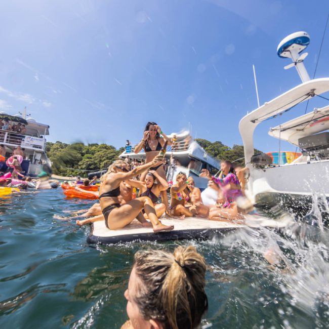 People on boats and floating inflatables enjoying a sunny day on the water, with some individuals splashing and playing near the boats during a lively Sydney boat party hire.