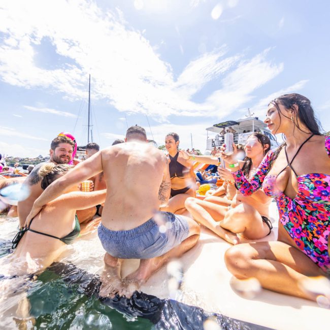 A group of people enjoying daylight on a catamaran party Sydney, some in swimwear, surrounded by water and other boats in the background.