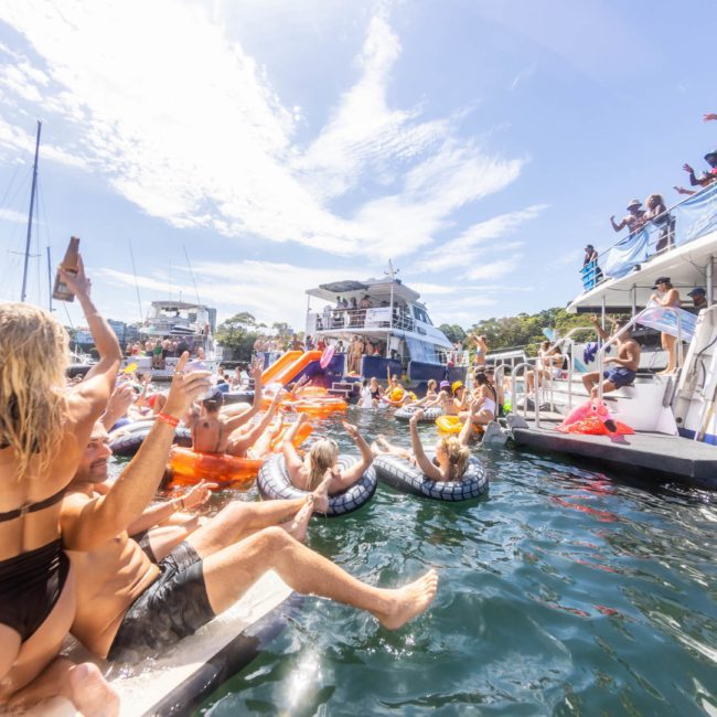 People enjoying a boat party, with many on pool floats and others on the boat, under a sunny sky. The atmosphere appears festive and lively, reminiscent of a DJ boat hire Sydney experience.