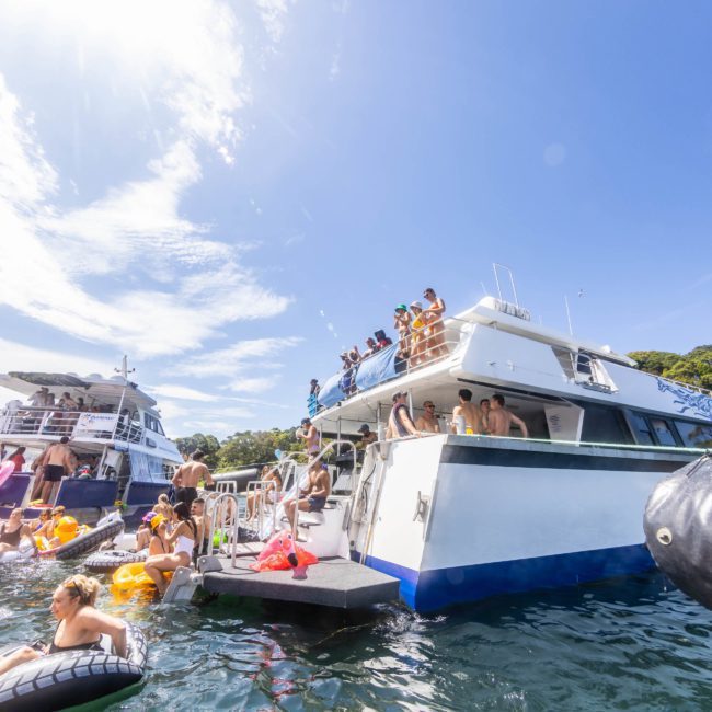 People enjoy a sunny day on and around a large white boat anchored in the water, with some swimming and using inflatable floats while others stand on the deck of their private yacht charter Sydney Harbour.