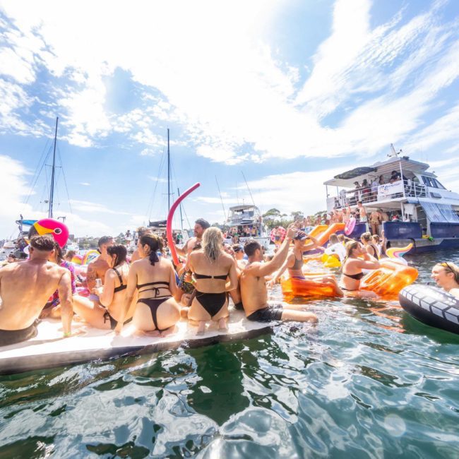 A group of people in swimsuits on floaties and paddleboards enjoys a sunny day in the water near boats, capturing the essence of a Private yacht charter Sydney Harbour.