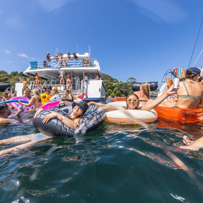 People enjoying a sunny day on the water with inflatable floats near a luxury yacht hire Sydney, some swimming, and others sitting on deck.