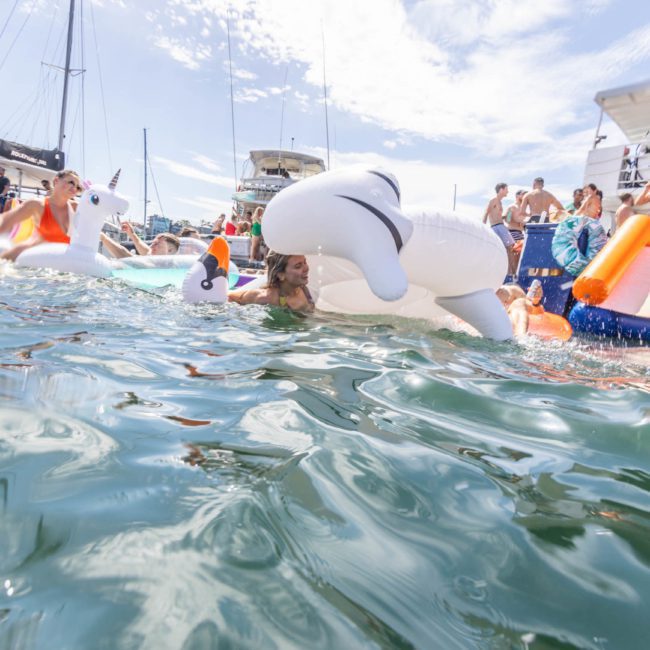 People are enjoying a sunny day on the water with inflatable toys, boats in the background, perfect for a Sydney boat party hire.