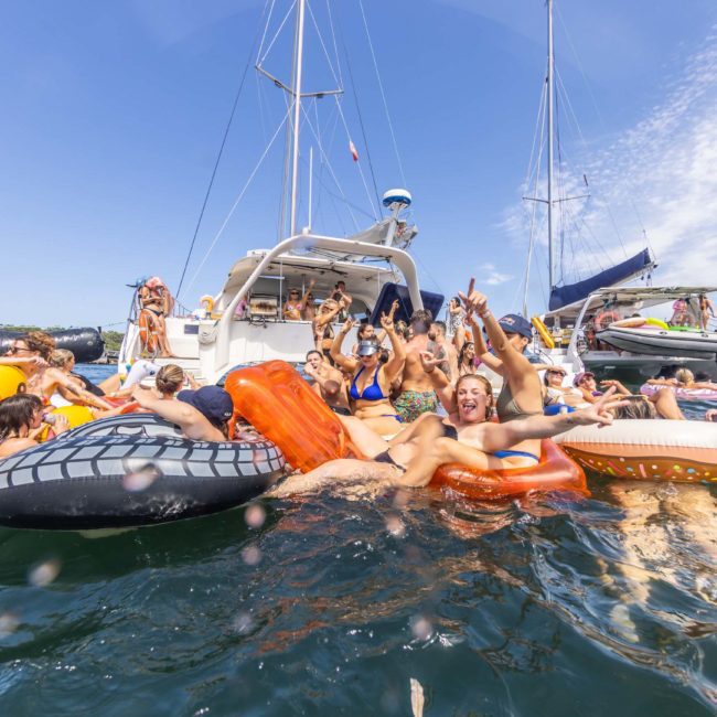 A group of people enjoying a sunny day on the water with inflatable rafts, surrounding a luxury yacht hire in Sydney.