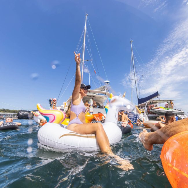 People in swimsuits enjoy a sunny day on the water with inflatable pool floats near anchored boats. One person in the center raises an arm, signaling a carefree atmosphere, perfect for those considering Sydney boat party hire.