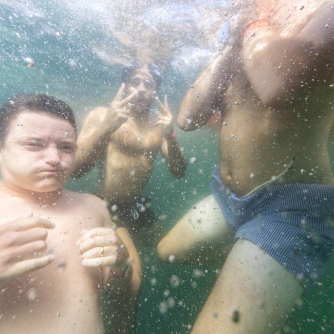 Three people are underwater in swimwear, with one showing a peace sign and others making facial expressions, surrounded by bubbles. Enjoy moments like these with a private yacht charter on Sydney Harbour.