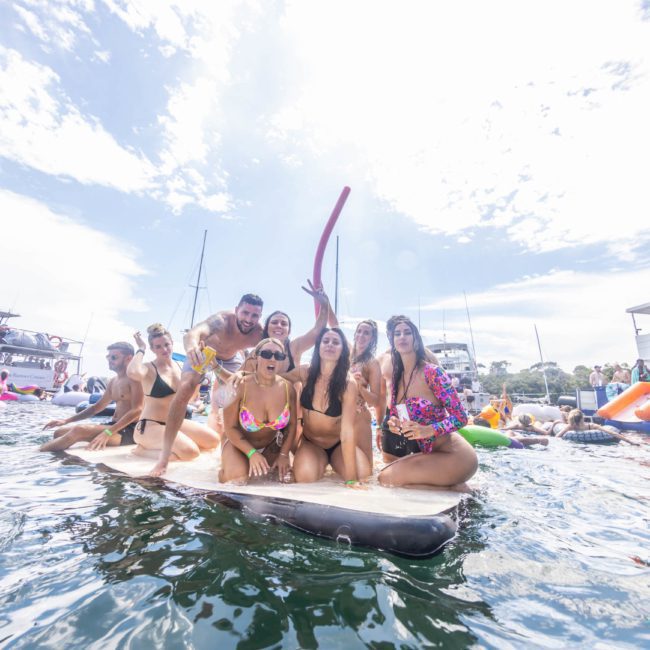 A group of people in swimwear are gathered on a floating platform in the water, holding a pool noodle, with other people and boats in the background on a sunny day during an exciting DJ boat hire Sydney event.