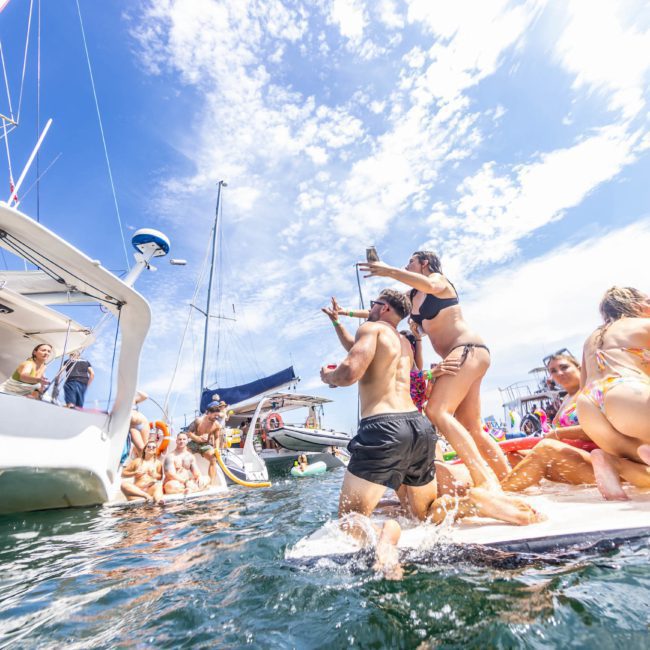 A group of people in swimwear enjoy a sunny day on and around boats, some standing on a floating dock beside the vessels, with a bright blue sky overhead. It's the perfect setting for a private yacht charter Sydney Harbour experience or a lively Sydney boat party hire event.