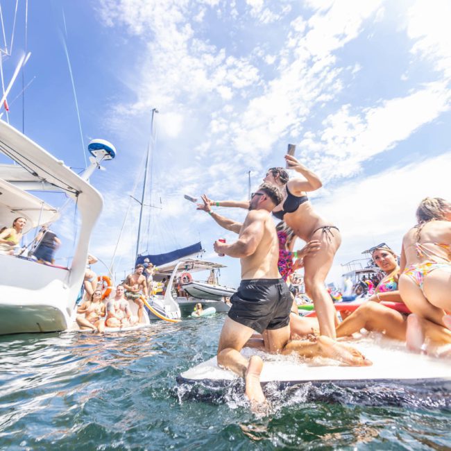 People are enjoying a lively party on a boat and in the water. The weather is sunny with a clear sky, and several boats are seen in the background. Some individuals are on a paddleboard, and others are swimming. This Sydney boat party hire scene perfectly captures fun moments under the sun.
