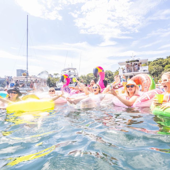 A group of people are floating on colorful inflatable pool toys in a sunny body of water surrounded by boats. They are smiling, wearing sunglasses, and appear to be enjoying themselves as part of a lively Sydney boat party hire event.