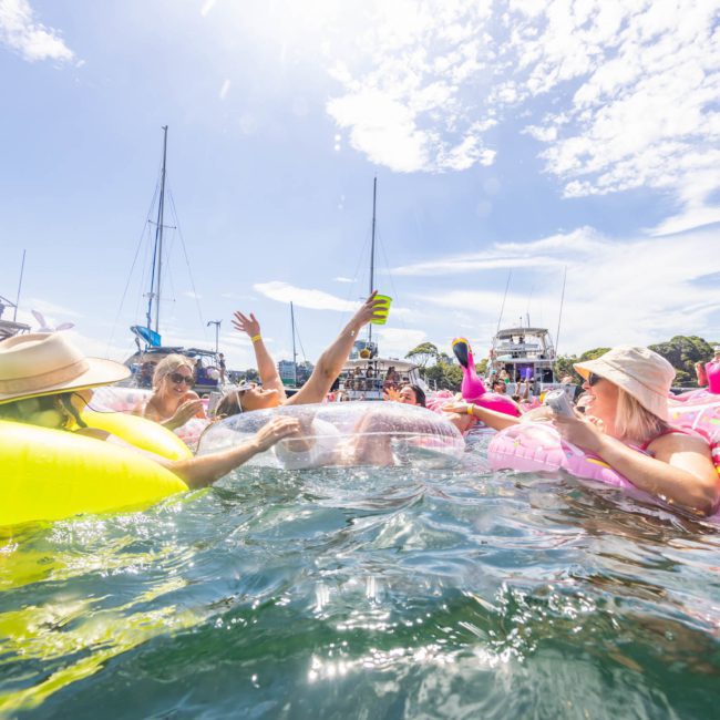 People are floating on inflatable rings in the water, surrounded by boats, enjoying a sunny day during a luxury yacht hire Sydney event.