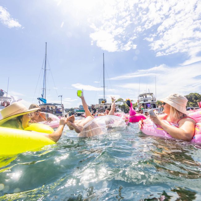 People enjoying a sunny day on colorful inflatable floats in the water, surrounded by boats and a clear sky, with a Catamaran party in Sydney adding to the fun.