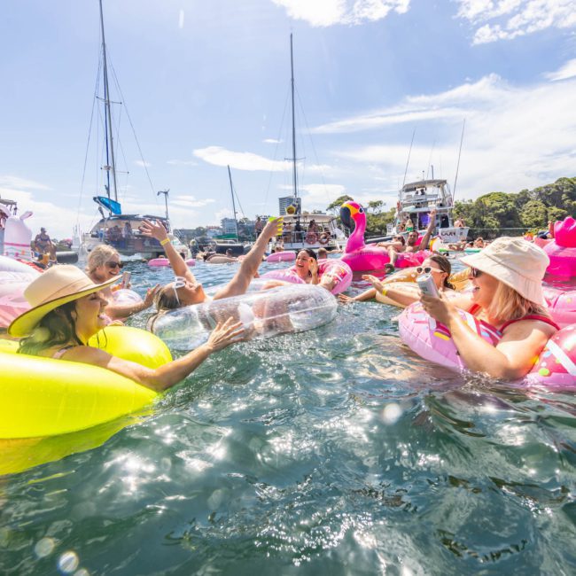 A group of people in colorful inflatable floaties enjoy a sunny day on the water, surrounded by anchored sailboats and yachts, perfect for a private yacht charter Sydney Harbour.