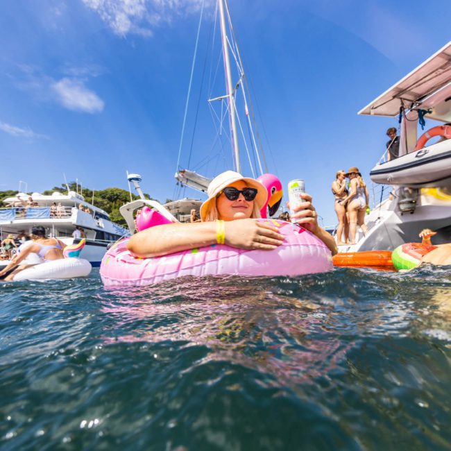 Person in a pink inflatable ring floating in water near boats, holding a phone and wearing sunglasses and a hat. Other people enjoying a Sydney boat party hire and inflatable items are also visible in the background.