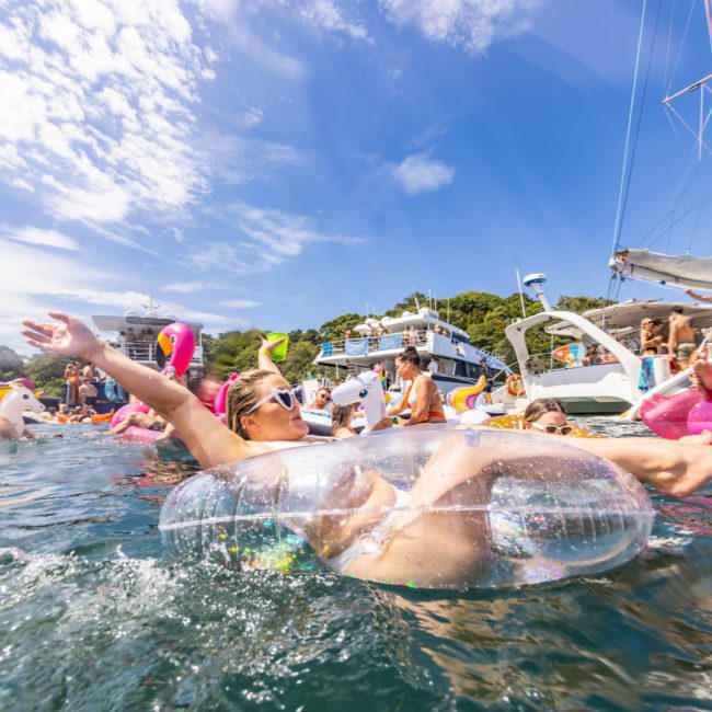 People are relaxing on inflatable floats in the water near several anchored boats on a sunny day, enjoying the ambiance of a luxury yacht hire Sydney.
