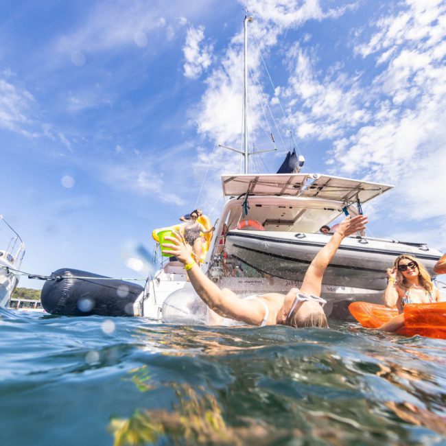 A group of people enjoying a sunny day on the water, some on a boat and others swimming nearby, perhaps part of a corporate boat event or private yacht charter in Sydney Harbour.
