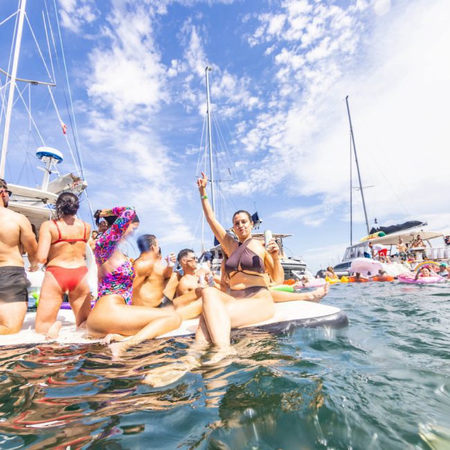 A group of people in swimsuits are relaxing on a large float in the water, surrounded by boats and other floating devices under a partly cloudy sky, enjoying what looks like an idyllic Sydney boat party hire.
