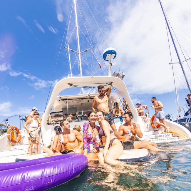 A group of people in swimwear enjoying time on and around a boat in clear, sunny weather. Some are in the water near an inflatable ring while others are on the boat during a lively Sydney boat party hire.