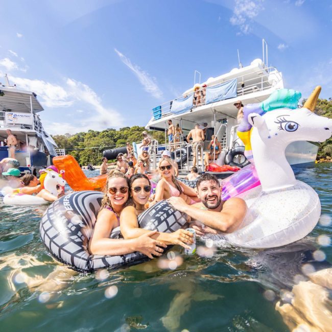 A group of people in water floats, including a unicorn float, enjoying themselves near a boat with more people on board under a clear sky. The scene sets the perfect vibe for a catamaran party in Sydney.