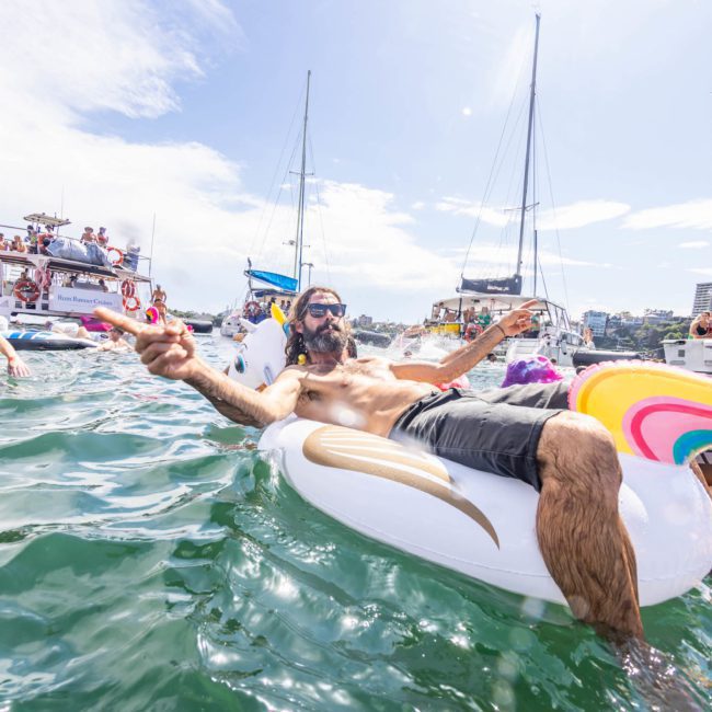 A man lounges on a unicorn float in the water, surrounded by boats and other people on floats, under a sunny sky, during a catamaran party in Sydney.