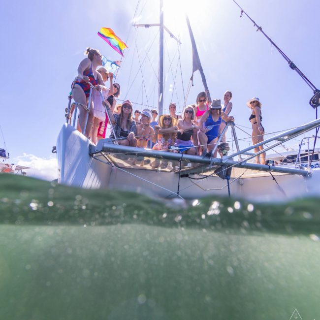 A group of people stands on the deck of a sailboat with a rainbow flag flying above them. The photo, taken from a low angle partly underwater, captures the essence of enjoying a private yacht charter on Sydney Harbour.