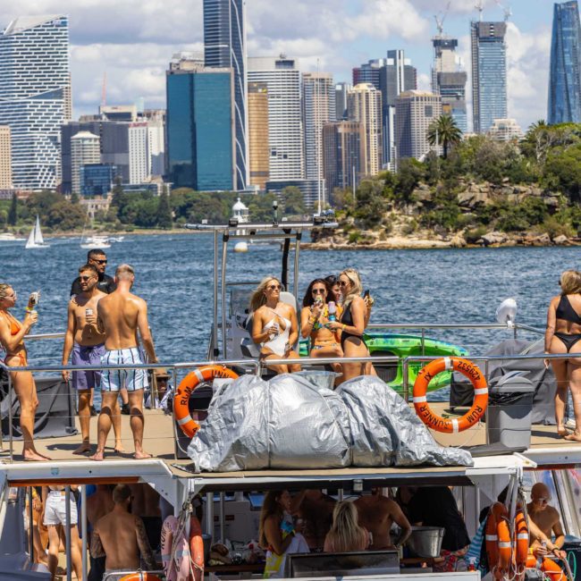 A group of people enjoy sunny weather on a luxury yacht with a city skyline in the background. Some are standing, some seated, and several are wearing swimsuits. The catamaran party is near a shoreline in Sydney Harbour.