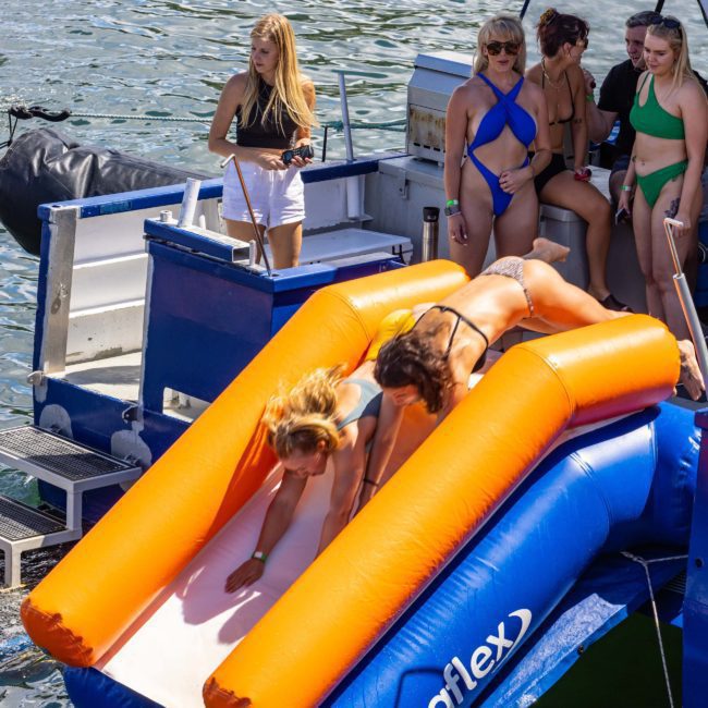 Two women in swimsuits slide down an inflatable slide into the water from a boat. Four others in swimsuits stand and sit on the boat, eagerly waiting their turn. Enjoy your adventure with a private yacht charter on Sydney Harbour or consider DJ boat hire for your next corporate event.
