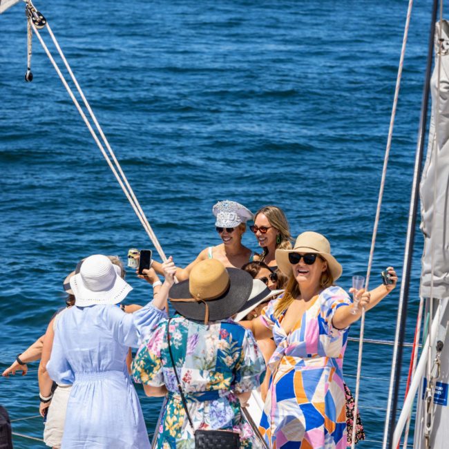 A group of people in colorful summer attire are standing on a catamaran, holding drinks and smiling, with the ocean in the background—a perfect scene for a Sydney boat party hire.