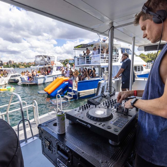 A DJ wearing a tank top and headphones mixes music on a boat, with a floating party of people and boats in the water in the background. It's a sunny day with scattered clouds, perfect for a catamaran party Sydney Harbour style.