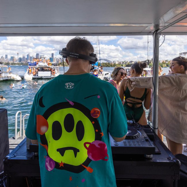A DJ in a green shirt with a smiley face design performs on a luxurious yacht. People are dancing and socializing around him, with other boats and the stunning Sydney Harbour skyline visible in the background.