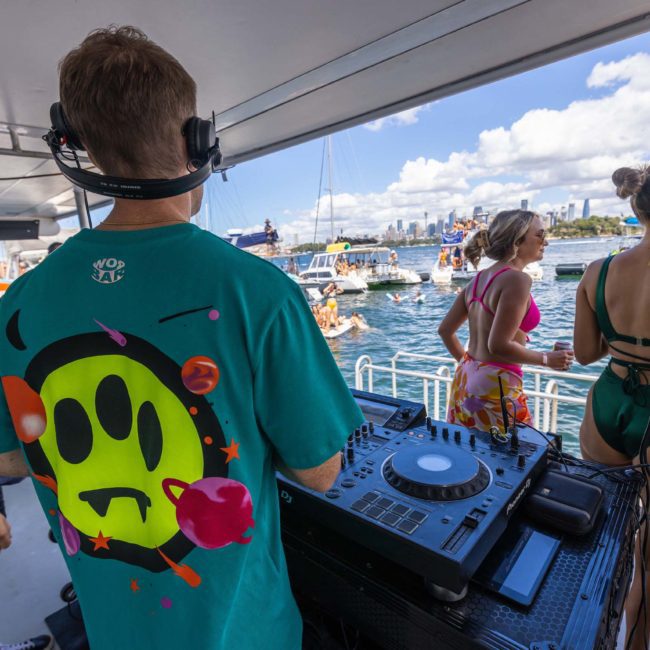 A DJ in a green shirt plays music on a private yacht charter in Sydney Harbour while people dance and socialize; other boats and the city skyline are visible in the background.