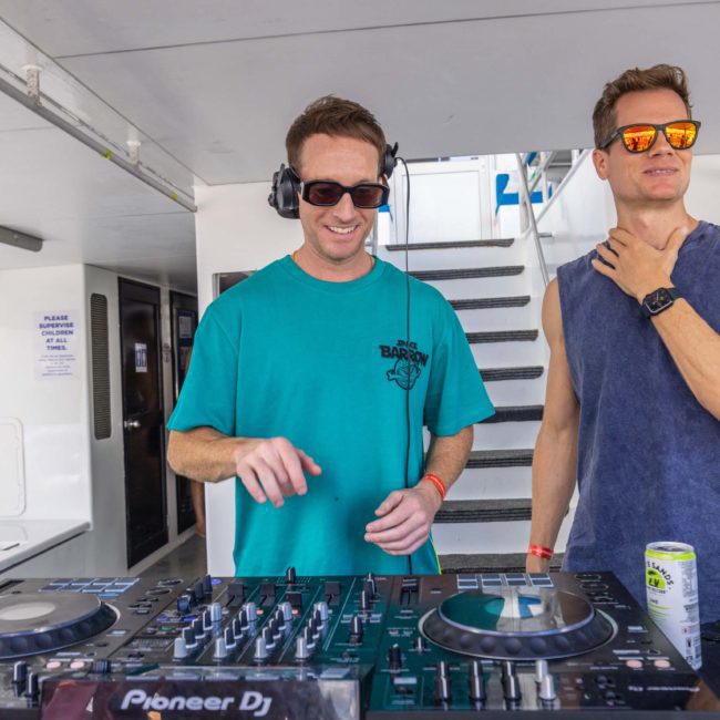 Two men are at a DJ booth on a boat. One is wearing headphones and adjusting controls on the console, while the other stands beside him, resting his hand on his neck. Experience this unique vibe with DJ boat hire Sydney for your next private yacht charter in Sydney Harbour.