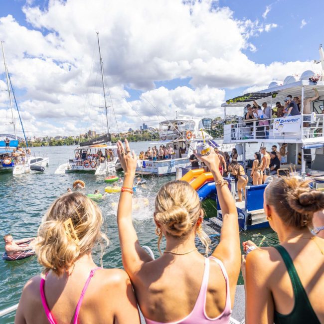 A group of people on a boat party, some standing and raising hands while others enjoy water activities. Several boats are anchored nearby under a partly cloudy sky. Perfect for those seeking a Catamaran party in Sydney.
