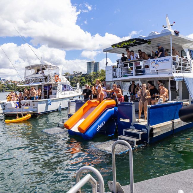 A group of people enjoying a sunny day on a boat with two orange slides leading into the water, other boats and a cityscape in the background, perfect for Sydney boat party hire or luxury yacht hire in Sydney.