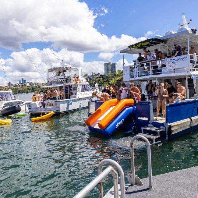 People are gathered on multiple boats, some using slides to enter the water. Kayaks and a paddleboard float nearby. The scene is set on a sunny day with a cityscape visible in the background, possibly during a Sydney boat party hire or Private yacht charter Sydney Harbour event.