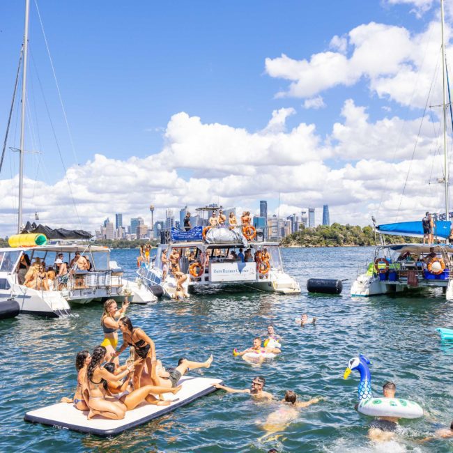 A group of people on boats and inflatables enjoy swimming and relaxing in a body of water with a city skyline visible in the background, reminiscent of a Sydney boat party hire.