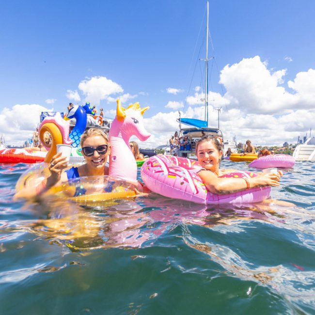 Two people float on inflatable pool toys in the water with boats in the background on a sunny day. The person on the left is holding a phone, and both are smiling, enjoying a relaxing moment during their catamaran party in Sydney.