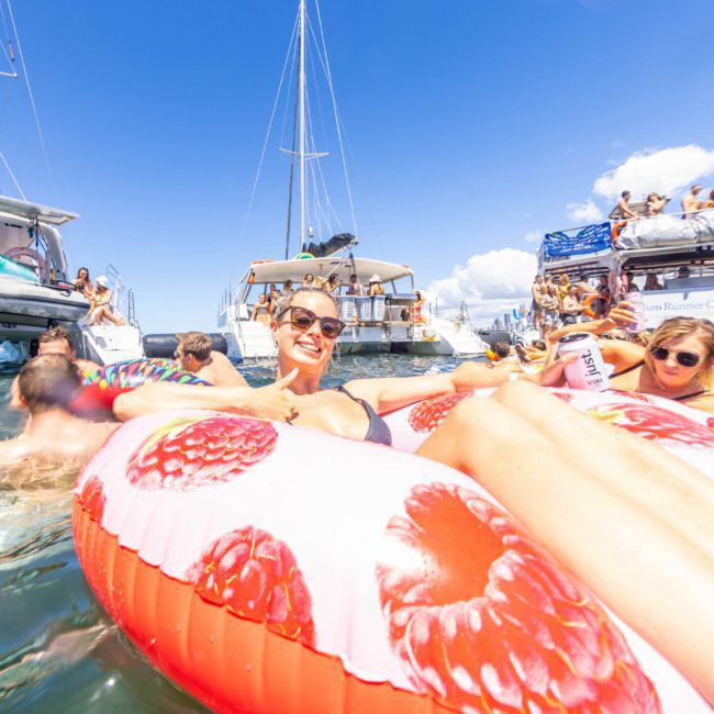 People relaxing on inflatable floats in the water near anchored boats under a clear blue sky, enjoying a catamaran party in Sydney.