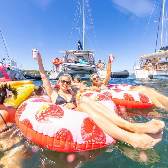 People relaxing on floaties, holding drinks, surrounded by boats on a sunny day. The water is clear, and the scene is festive with options for a private yacht charter Sydney Harbour in the background.