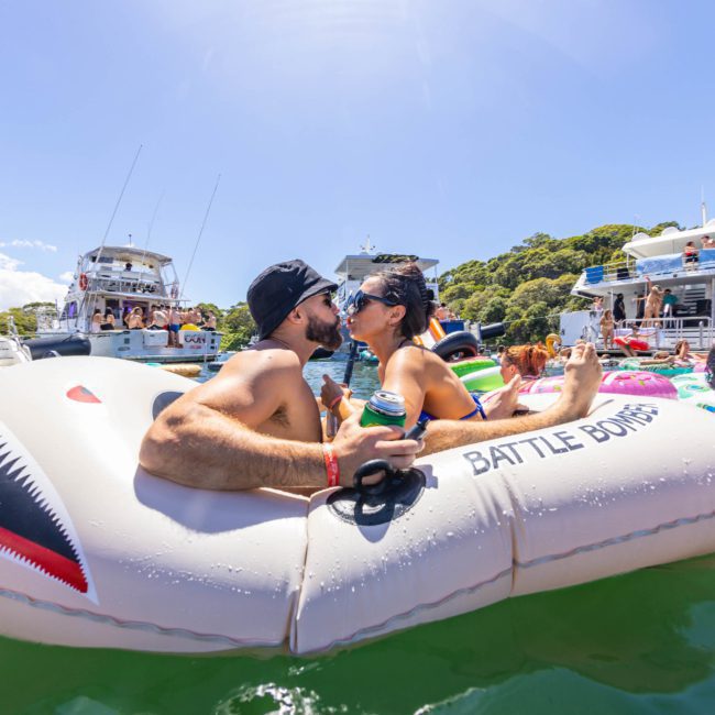 A man and woman share a kiss while lounging on a shark-shaped inflatable raft with "BATTLE BOATS" printed on it, surrounded by others on various inflatables in a bustling water scene, accentuating the lively atmosphere often seen around Sydney Harbour's luxury yacht hires.