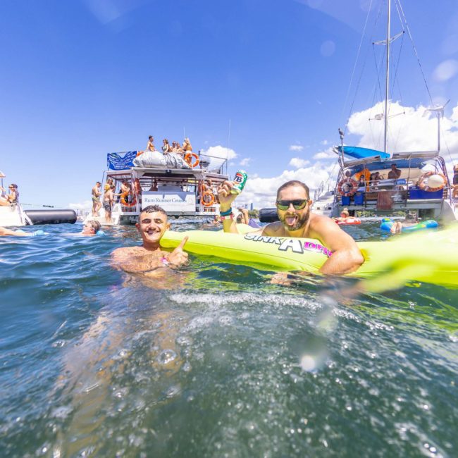 Two people in the foreground float on an inflatable in the ocean. In the background, groups of people socialize and relax on anchored boats under a clear blue sky, embodying the essence of a Sydney boat party hire event.