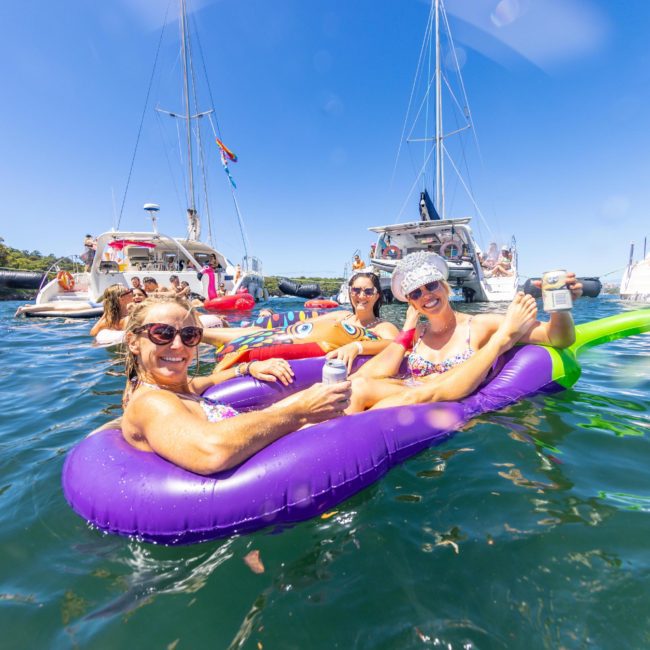 Three people are relaxing on inflatable floats in the water near anchored boats on a sunny day, enjoying a private yacht charter Sydney Harbour.